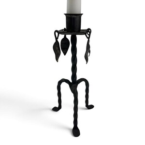 May include: A black metal candlestick holder featuring a twisted central stem and three matching twisted legs. The top platform includes a cylindrical candle cup and is decorated with four hanging metal leaf-shaped ornaments.