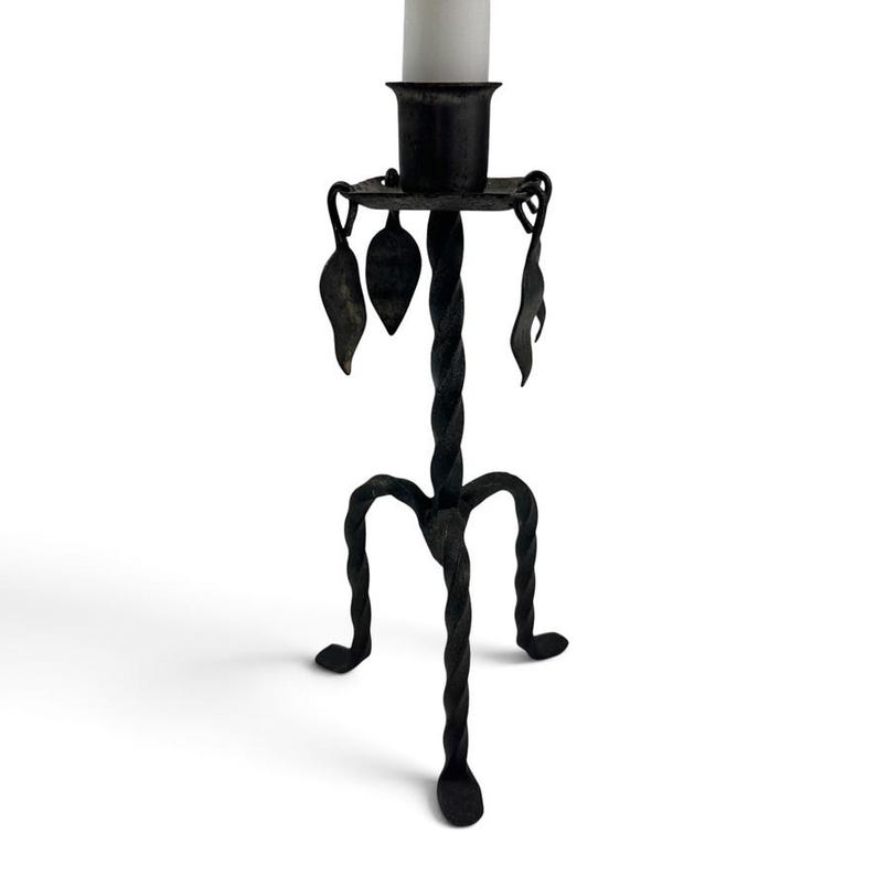 May include: A black metal candlestick holder featuring a twisted central stem and three matching twisted legs. The top platform includes a cylindrical candle cup and is decorated with four hanging metal leaf-shaped ornaments.