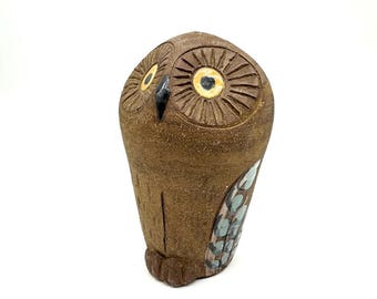 May include: A brown ceramic owl figurine with detailed carved feather and eye patterns. The owl has yellow and black eyes and a small beak. The wings are decorated with blue and green accents. The owl is standing upright.