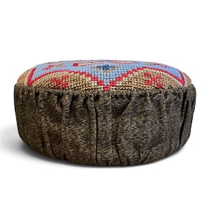 May include: A round floor cushion featuring a top panel with a cross-stitch pattern in light blue, tan, and red. The sides of the cushion are made of a dark brown, textured fabric with vertical pleats running around the entire circumference.