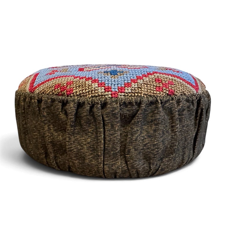 May include: A round floor cushion featuring a top panel with a cross-stitch pattern in light blue, tan, and red. The sides of the cushion are made of a dark brown, textured fabric with vertical pleats running around the entire circumference.