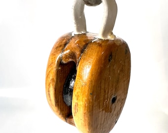 May include: A vintage wooden pulley block featuring a polished light brown wood finish, a central dark metal wheel, and a thick white painted metal hook attachment at the top. The pulley exhibits a smooth, rounded shape with visible wood grain.