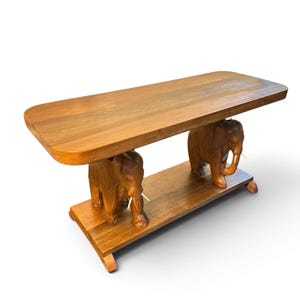 Puede incluir: Una mesa rectangular de madera con una superficie superior lisa sostenida por dos figuras de elefantes talladas que se apoyan sobre una base de madera a juego. Los elefantes están hechos de madera marrón e incluyen colmillos claros, creando una estructura robusta.