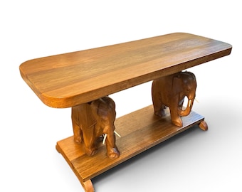 Op de afbeelding: Een rechthoekige houten tafel met een glad oppervlak dat wordt ondersteund door twee uitgesneden olifantenfiguren die op een bijpassende houten basis staan. De olifanten zijn gemaakt van bruin hout en hebben lichtgekleurde slagtanden, wat een stevige structuur vormt.