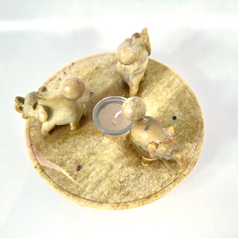 May include: A beige stone candle holder with three small elephant figurines and a small candle. The elephants are arranged around the edge of a circular base. The candle is in a small metal container.