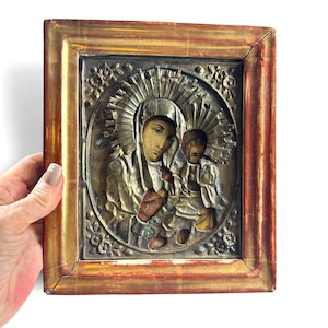 May include: A rectangular religious icon featuring a metallic relief of a figure holding a child, surrounded by a sunburst halo. The central panel is encased in a weathered, distressed wooden frame with a gold and reddish-brown finish.