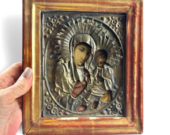 May include: A rectangular religious icon featuring a metallic relief of a figure holding a child, surrounded by a sunburst halo. The central panel is encased in a weathered, distressed wooden frame with a gold and reddish-brown finish.