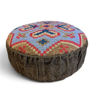 May include: A round pincushion featuring a top panel with a cross-stitch pattern in light blue, red, dark blue, and tan. The sides are made of a dark brown, textured fabric with a gathered, pleated design around the circumference.