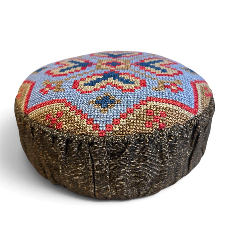 May include: A round pincushion featuring a top panel with a cross-stitch pattern in light blue, red, dark blue, and tan. The sides are made of a dark brown, textured fabric with a gathered, pleated design around the circumference.