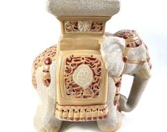 May include: A ceramic elephant-shaped garden stool featuring a textured beige finish with smooth, glossy red and tan decorative accents. The stool has a flat, rectangular top and intricate cut-out patterns along the sides and back.