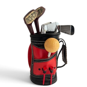 Op de afbeelding: Een miniatuur golftas van rood en zwart kunstleer met witte stiksels. De tas bevat twee bruine putters met een rasterpatroon op het slagvlak, een zilverkleurige ijzeren club, een gele golfbal en twee kleine rode golftees op de voorzak.