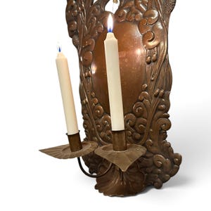K&ouml;nnte beinhalten: Ein Wandkerzenhalter aus Kupfer mit einer verzierten, gepr&auml;gten R&uuml;ckplatte mit Blumen- und Schn&ouml;rkelmotiven. Zwei geschwungene Arme ragen nach vorne, jeder mit einem geriffelten Tropfteller und einer brennenden, cremewei&szlig;en Stabkerze.