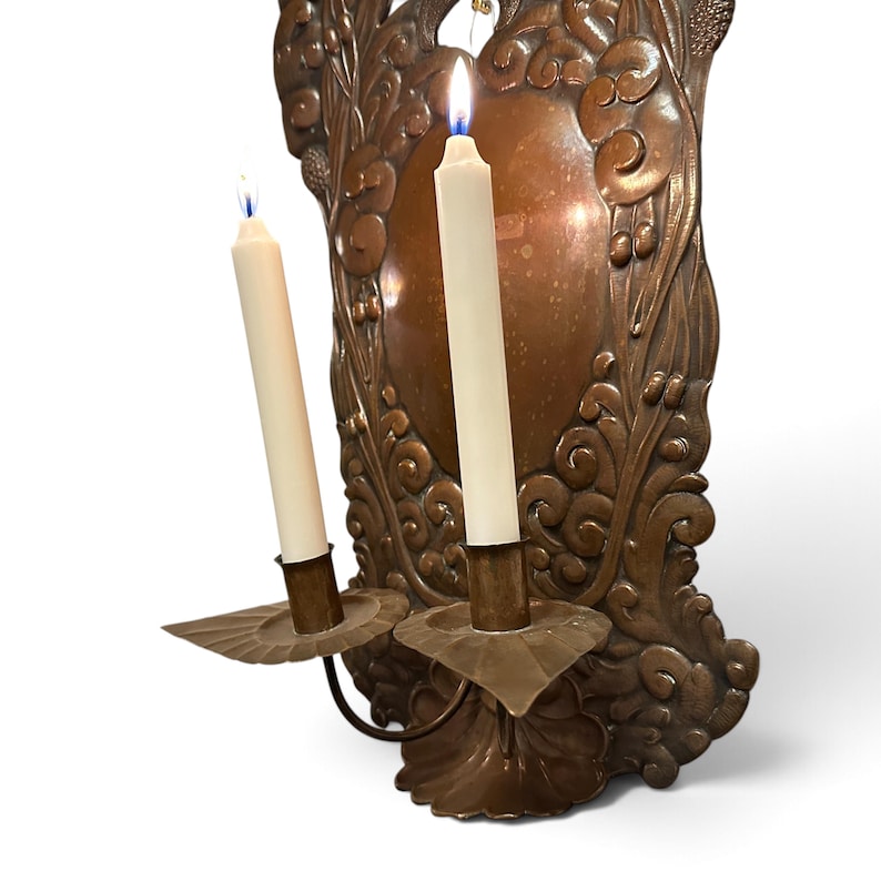 K&ouml;nnte beinhalten: Ein Wandkerzenhalter aus Kupfer mit einer verzierten, gepr&auml;gten R&uuml;ckplatte mit Blumen- und Schn&ouml;rkelmotiven. Zwei geschwungene Arme ragen nach vorne, jeder mit einem geriffelten Tropfteller und einer brennenden, cremewei&szlig;en Stabkerze.