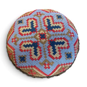 May include: A round, stuffed textile cushion featuring a detailed cross-stitch pattern in light blue, dark blue, red, and tan thread. The geometric design radiates from the center, creating a symmetrical, intricate, and colorful needlepoint motif.