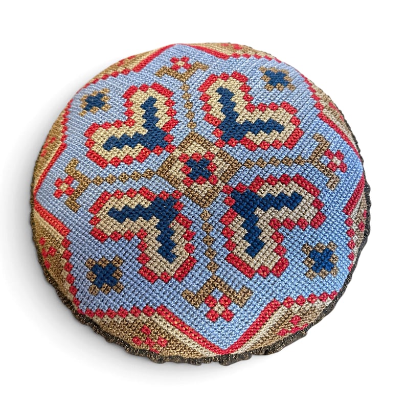 May include: A round, stuffed textile cushion featuring a detailed cross-stitch pattern in light blue, dark blue, red, and tan thread. The geometric design radiates from the center, creating a symmetrical, intricate, and colorful needlepoint motif.