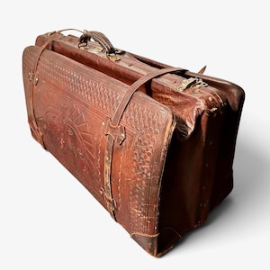 May include: A rectangular brown leather suitcase featuring an embossed sun design on the front panel, a textured geometric border, and two vertical leather straps with metal buckles. The luggage includes a top handle and metal hardware accents.