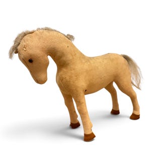 May include: A tan-colored plush horse toy with a textured fabric body, a light-colored synthetic mane and tail, and dark brown fabric hooves. The horse has a downward-facing head position, small dark eyes, and visible stitched seams along the neck.