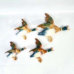 May include: A set of four ceramic mallard duck wall plaques in varying sizes, each depicted in flight with wings spread. The ducks feature glossy finishes in shades of green, brown, white, and blue, with detailed moulded feathers and open beaks.