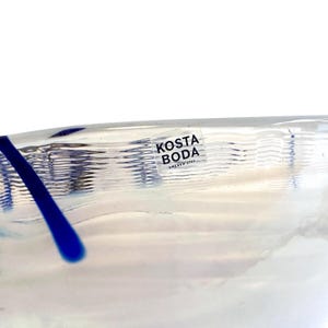 Pu&ograve; includere: Una ciotola in vetro trasparente caratterizzata da una superficie testurizzata e ondulata con una linea di accento in vetro blu. Un adesivo quadrato &egrave; applicato sul lato della ciotola con il testo nero KOSTA BODA SWEDEN 1742, indicando il produttore.