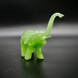 K&ouml;nnte beinhalten: Eine durchscheinende, hellgr&uuml;ne Elefantenfigur mit erhobenem R&uuml;ssel. Der Elefant hat eine glatte, gl&auml;nzende Oberfl&auml;che und steht vor einem dunklen Hintergrund. Die Figur ist ein dekoratives Objekt.