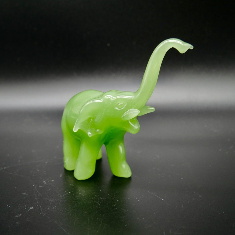 K&ouml;nnte beinhalten: Eine durchscheinende, hellgr&uuml;ne Elefantenfigur mit erhobenem R&uuml;ssel. Der Elefant hat eine glatte, gl&auml;nzende Oberfl&auml;che und steht vor einem dunklen Hintergrund. Die Figur ist ein dekoratives Objekt.