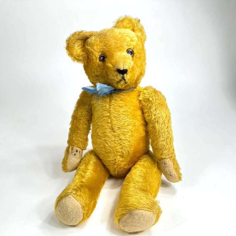 May include: A vintage golden yellow teddy bear with a light blue bow tie. The bear has black eyes, a black nose, and tan-colored paws. The plush toy is sitting upright, showcasing its classic design.