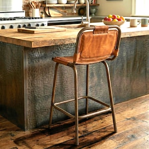 May include: A brown leather and metal bar stool with a rectangular back and seat. The stool has a metal frame with a footrest and is positioned in front of a wooden countertop.