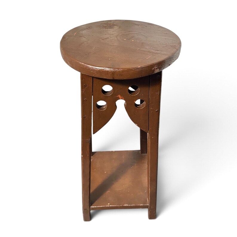 May include: A brown wooden plant stand featuring a circular top, four straight vertical legs, and a square lower shelf. The upper section includes decorative cutouts with circular and pointed arch shapes on each of the four sides.