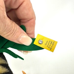 May include: A green felt wing of a stuffed animal is held by a hand. A yellow tag is attached with a silver button. The tag reads "Outer covering 51% wool 49% cotton."