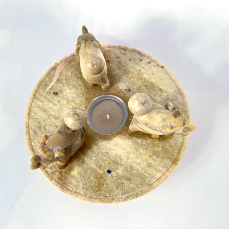May include: A round, beige candle holder with three small animal figurines and a central candle. The figurines are light brown and cream-colored, and the candle is in a silver metal holder. The candle holder has a speckled pattern.