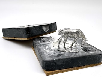 May include: A silver-colored metal horse figurine standing on a black rectangular mold base with a cork-like bottom layer. The mold is split into two pieces, with one half showing the negative impression of the horse and the other half open.