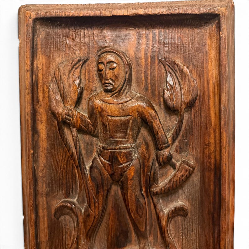 May include: A rectangular brown wooden wall plaque featuring a hand-carved relief of a figure wearing a hooded garment. The figure holds a long staff in one hand and a curved horn in the other, surrounded by stylized leaf motifs.