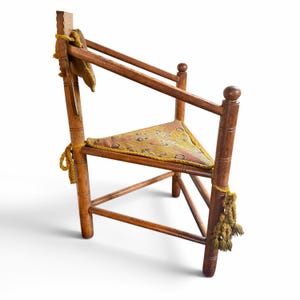 May include: A wooden corner chair with a triangular seat featuring a patterned yellow and multi-colored fabric cushion. The chair has cylindrical legs, a high backrest with a small attached cushion, and decorative yellow tassels tied to the frame.