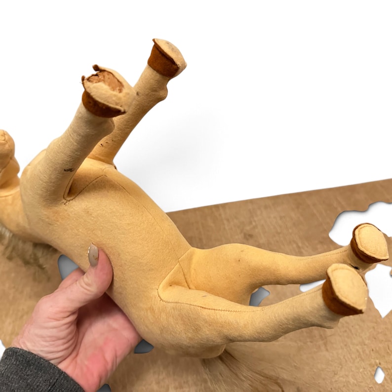 May include: A tan plush horse toy with a light-colored mane and tail. The horse features brown fabric hooves at the end of each leg. One of the front hooves shows visible wear and tear with exposed material at the tip of the hoof area.
