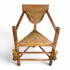 May include: A wooden triangular chair featuring a carved backrest with a geometric pattern. The seat and back cushion are upholstered in a fabric with a moth-like print in shades of pink, yellow, and brown, finished with yellow braided trim and tassels.
