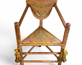 Op de afbeelding: Een driehoekige houten stoel met een uitgesneden rugleuning met geometrisch patroon. De zitting en het rugkussen zijn bekleed met een stof met een mot-achtig patroon in roze, gele en bruine tinten, afgewerkt met gele gevlochten randen en kwastjes.
