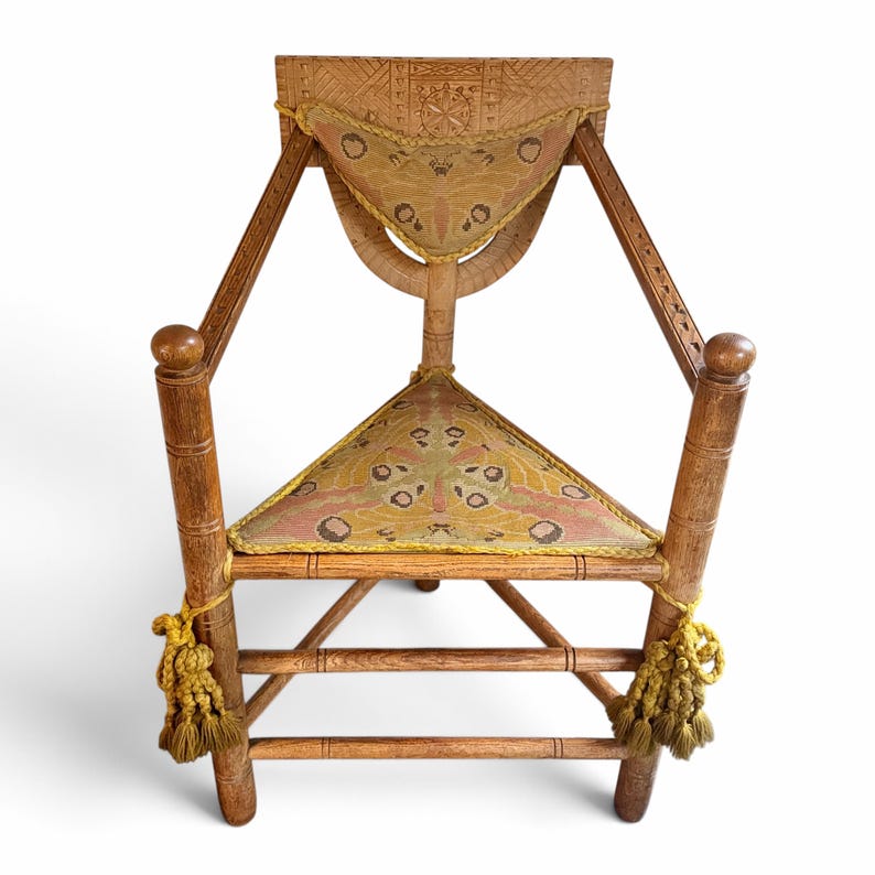 May include: A wooden triangular chair featuring a carved backrest with a geometric pattern. The seat and back cushion are upholstered in a fabric with a moth-like print in shades of pink, yellow, and brown, finished with yellow braided trim and tassels.