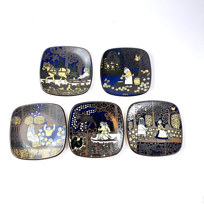 Op de afbeelding: Vijf vierkante decoratieve borden met afgeronde hoeken. Elk bord heeft een andere sc&egrave;ne met figuren, bomen en dieren in een donkerblauw, goud en wit kleurenpalet. De borden hebben een vintage uitstraling.
