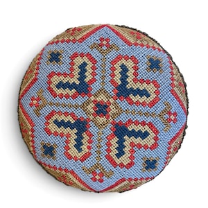 May include: A round, stuffed textile cushion featuring a detailed cross-stitch pattern. The design displays a central diamond motif in shades of light blue, dark blue, red, and tan, arranged in a symmetrical, geometric, repeating pattern.