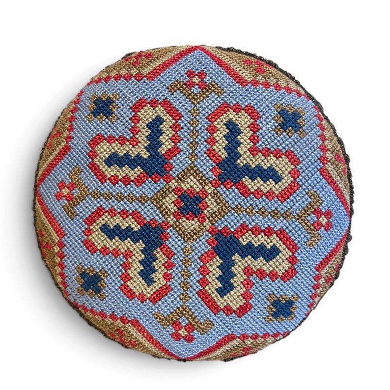 May include: A round, stuffed textile cushion featuring a detailed cross-stitch pattern. The design displays a central diamond motif in shades of light blue, dark blue, red, and tan, arranged in a symmetrical, geometric, repeating pattern.