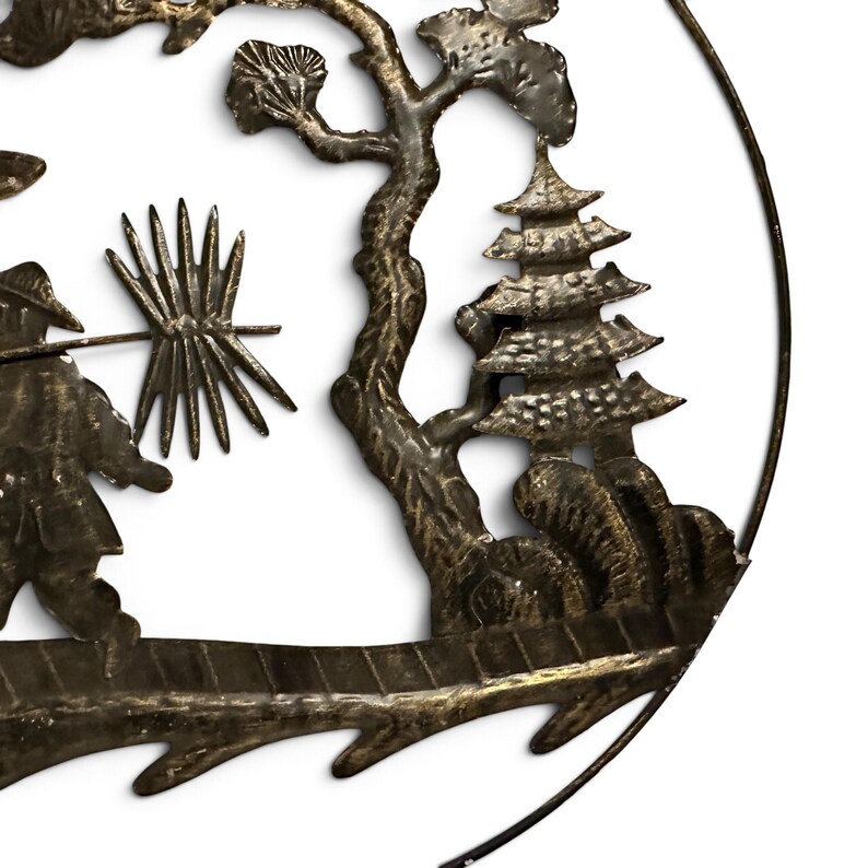 May include: A circular metal wall art piece featuring a dark bronze finish with a textured surface. The design depicts a figure carrying a spiky object on a pole, a gnarled tree, and a multi-tiered pagoda structure, all enclosed within a thin metal frame.