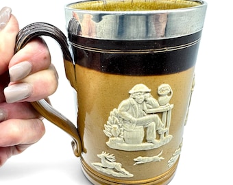 Puede incluir: Una taza de cerámica de color tostado y marrón oscuro con un borde de metal plateado y un asa marrón oscuro. El exterior está decorado con figuras en relieve blanco, incluyendo una persona sentada en un barril y animales.