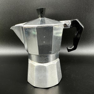 Puede incluir: Una cafetera de aluminio plateado con un mango de pl&aacute;stico negro. La cafetera tiene un cuerpo redondeado con un pico y una tapa.