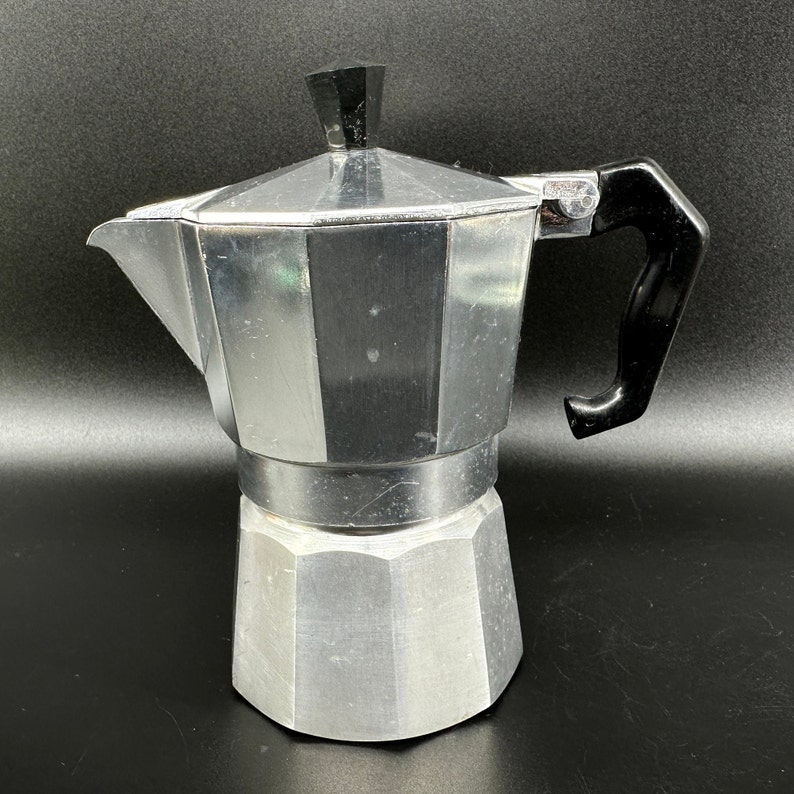 Puede incluir: Una cafetera de aluminio plateado con un mango de pl&aacute;stico negro. La cafetera tiene un cuerpo redondeado con un pico y una tapa.