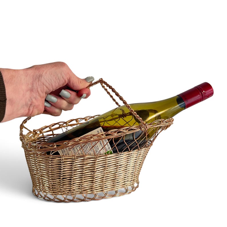 Puede incluir: Un portabotellas de vino de alambre en tono dorado con base ovalada y patr&oacute;n de metal tejido. El soporte cuenta con un asa de alambre trenzado y est&aacute; dise&ntilde;ado para sostener una sola botella de vino en &aacute;ngulo, mostrando el cuello y la etiqueta.