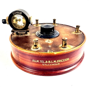 Op de afbeelding: Een ronde houten radiocomponent met een donkerbruine afwerking, voorzien van de tekst ALLM. TEL. A. B. L. M. ERICSSON STOCKHOLM. Het bevat een zwarte centrale draaiknop, vier messing aansluitpunten en een zwarte ronde SONAR MOD. 1926/27 D.R.G.M. meter.
