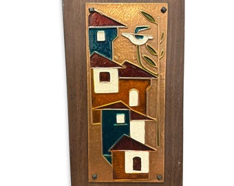 May include: A rectangular wall plaque featuring a central copper panel mounted on a dark wood base. The copper panel displays an enameled scene of stacked houses in shades of blue, brown, and cream, with a stylized bird perched on a branch.