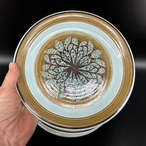 Puede incluir: Una pila de platos de cerámica con un diseño floral. Cada plato presenta un centro azul claro, un borde marrón y un patrón floral marrón y turquesa en el centro. Los platos son sostenidos por una mano sobre un fondo negro.