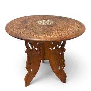 May include: A round wooden side table featuring intricate floral carvings across the surface and a central circular inlay depicting a tree. The base consists of two interlocking panels with openwork cutouts, finished in a warm brown tone.