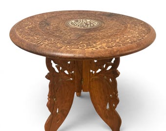 May include: A round wooden side table featuring intricate floral carvings across the surface and a central circular inlay depicting a tree. The base consists of two interlocking panels with openwork cutouts, finished in a warm brown tone.